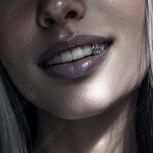 May include: Close-up of a person's mouth with a silver tooth gem. The gem is a cross design. The person has pale skin, light hair, and dark purple lipstick. The lips are glossy.