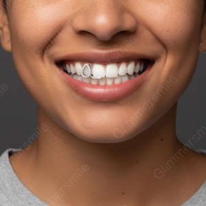 May include: A close-up of a person smiling, displaying a silver tooth gem. The gem is shaped like the letter "O" and is adorned with small, sparkling stones. The person's teeth are white and evenly spaced.