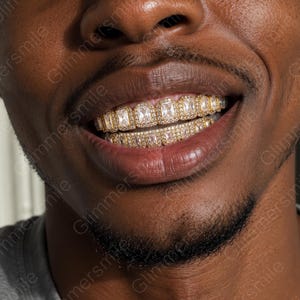 May include: A set of gold-tone dental grills featuring rectangular-cut clear stones set in a row on both the upper and lower teeth. The design includes a border of smaller pavé-style stones surrounding each larger central rectangular stone.