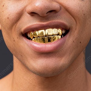 May include: Close-up of a person smiling, showcasing a full set of gold-colored teeth grillz. The grillz are highly reflective, catching the light and creating a bright, bold appearance. The teeth are perfectly aligned, and the gold color is consistent.