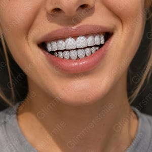 Op de afbeelding: Een set zilverkleurige tandengrills die zowel de boven- als ondertanden bedekken. Elke individuele tandkap is dicht bezet met talloze kleine, heldere, reflecterende steentjes die zorgen voor een sterk getextureerd en sprankelend uiterlijk.