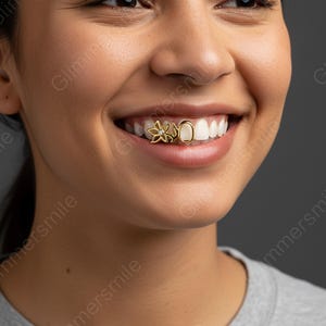 May include: A person smiling, displaying gold tooth jewellery. The jewellery features a flower and an oval design, both in gold. The person is wearing a grey top.