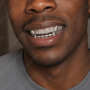 Könnte beinhalten: Ein silberfarbenes Grillz-Set für die oberen und unteren Zähne mit metallischem Finish und spitz zulaufenden Eckzahn-Designs. Das Zahnschmuck-Accessoire bedeckt die Vorderzähne und zeichnet sich durch eine glatte, reflektierende Oberfläche aus.