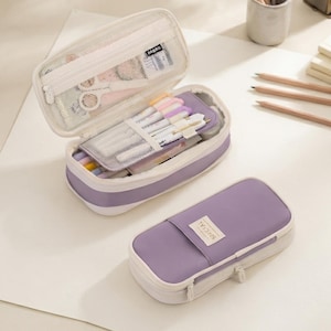 Pode incluir: Um estojo de lápis lavanda e creme com fecho de correr. Um estojo está aberto, revelando um bolso de rede e uma aba central com elásticos para canetas. O outro estojo está fechado e apresenta uma pequena etiqueta retangular com o texto SPECIAL.