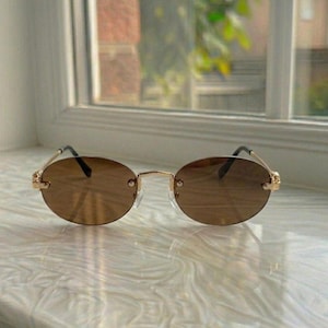 May include: Rimless oval sunglasses featuring brown tinted lenses and a thin gold-tone metal frame. The design includes slender gold temples and a delicate bridge connecting the two lenses, providing a lightweight and streamlined appearance.