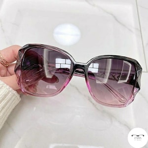 May include: Oversized sunglasses featuring a black to pink gradient frame with faceted edges. The lenses have a dark purple to light pink gradient tint. The temples display a geometric cutout pattern. The text ZARIX FINGS is printed in the corner.