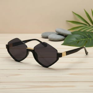 Puede incluir: Gafas de sol negras con montura geométrica en forma de diamante y lentes oscuras. Las patillas son negras con detalles dorados cerca de las bisagras. Las gafas de sol están sobre una superficie de madera clara.