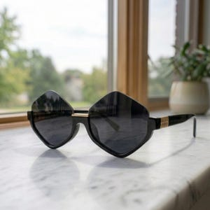 Könnte beinhalten: Schwarze Sonnenbrille mit einem markanten, geometrischen, rautenförmigen Rahmendesign. Die Brille verfügt über dunkel getönte Gläser, einen dünnen goldfarbenen Steg zwischen den Gläsern und goldfarbene gerippte Details an den Bügeln.