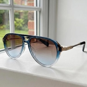 May include: Aviator sunglasses featuring a translucent blue gradient frame and brown tinted lenses. The temples are constructed from gold-toned metal with ribbed detailing and black acetate tips, providing a structured look for these eyewear frames.