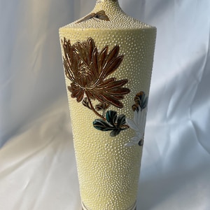 May include: A tall, tapered ceramic vase with a pale yellow, textured surface featuring raised white dots. The vase is decorated with a large, metallic bronze-colored chrysanthemum flower, green leaves, and a small butterfly near the narrow neck.