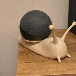 May include: A dark gray smart speaker sits atop a cream-colored snail-shaped stand. The stand has two antennae and a rounded body, giving it a whimsical appearance. The speaker is a rounded, fabric-covered device.