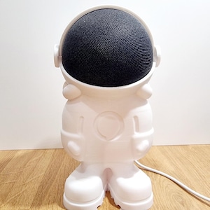 May include: A white astronaut-shaped speaker stand with a black mesh speaker. The stand has a rounded helmet, arms, legs, and a circular design on the chest. A white power cord extends from the back. The speaker is designed to look like an astronaut.
