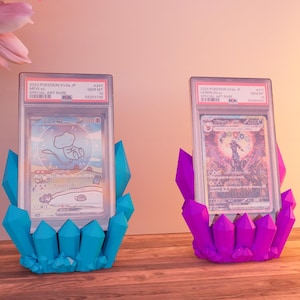 Op de afbeelding: Twee Pokemon-verzamelkaarten in beschermende hoesjes, tentoongesteld op blauwe en paarse kristalvormige standaards. De kaarten bevatten kleurrijke illustraties en Japanse tekst.