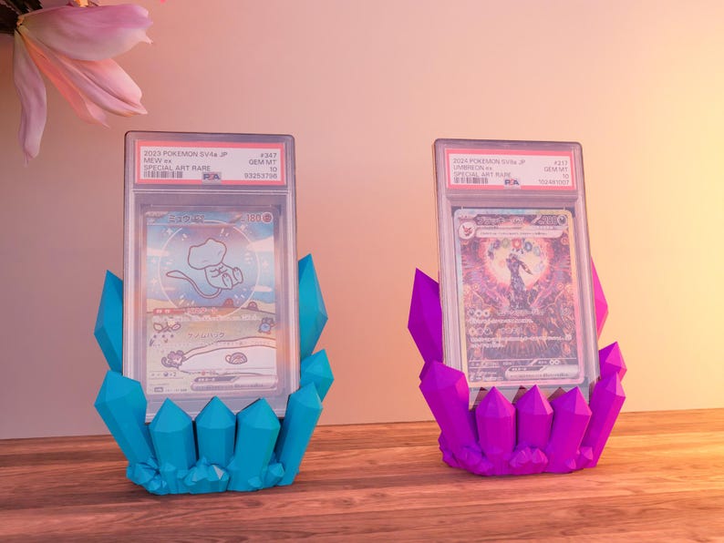 Op de afbeelding: Twee Pokemon-verzamelkaarten in beschermende hoesjes, tentoongesteld op blauwe en paarse kristalvormige standaards. De kaarten bevatten kleurrijke illustraties en Japanse tekst.
