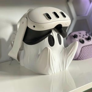 May include: A white VR headset rests on a ghost-shaped stand. The headset has a black visor and three dark lenses. The stand is white with two black eye-shaped cutouts. A purple game controller is in the background.