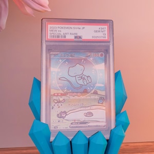 Op de afbeelding: Een beoordeelde Pok&eacute;mon-kaart met Mew ex, weergegeven in een doorzichtige hoes. De kaart wordt ondersteund door een levendige blauwe kristalvormige standaard. De kaartdetails bevatten tekst in het Japans en Engels.