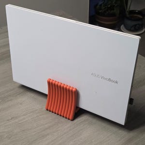 May include: A white ASUS VivoBook laptop is propped up by an orange stand on a gray wooden surface. The laptop is closed, displaying the ASUS VivoBook logo. The stand is made of vertical, curved orange bars.