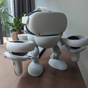 May include: A white VR headset and controllers are displayed on a gray stand. The stand is designed to resemble a robot. The VR headset is on top, with the controllers held in place by the stand's arms.