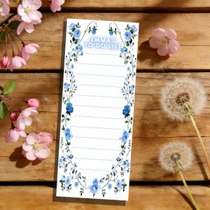 May include: A white to-do list notepad with a blue floral design. The notepad has a blue floral border and the text "EMMA'S TO DO LIST" at the top. It rests on a wooden surface, with pink flowers and dandelions.