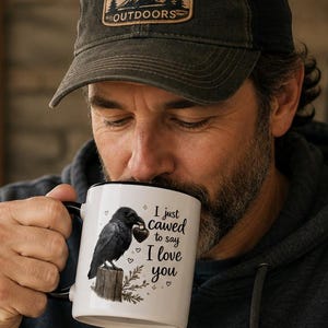 May include: White ceramic mug with a black rim and handle, featuring a black raven illustration and the text "I just cawed to say I love you." The mug is displayed with a "Best Dad Ever" sign in the background.