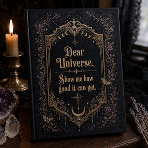 Op de afbeelding: Een zwart notitieboek met harde kaft en gouden letters met de tekst Dear Universe, Show me how good it can get. De kaft is versierd met een sierlijk gouden kader, paarse bloemmotieven, vlinders en een symbool van een halve maan.
