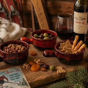 May include: Three round ceramic bowls with handles, featuring a deep red and black drip glaze. The bowls contain walnuts, olives, and crackers. A bottle of red wine labelled Après ski, two glasses of wine, and a card with a skier and the text Après ski, good food, good friends, good times are present.