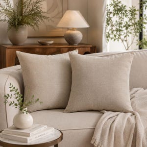 May include: Two square throw pillows in a solid light beige color with a visible woven fabric texture. The pillows are positioned side by side on a light-colored sofa, featuring a simple design with clean edges and a consistent neutral tone.