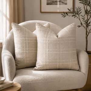 May include: Two square throw pillows in a light beige color featuring a subtle white grid pattern. The pillows are made of a textured fabric and are positioned upright, side by side, displaying a clean, structured design with visible stitching.