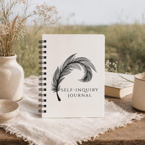 Puede incluir: Un diario de espiral blanco con una ilustración de una pluma negra y el texto "SELF-INQUIRY JOURNAL" impreso en la portada. El cuaderno cuenta con una encuadernación de espiral metálica negra en el lado izquierdo y una cubierta de papel blanco roto.