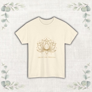 Puede incluir: Camiseta color crema con un diseño de flor de loto dorada y el texto "TRUST THE PROCESS" debajo. El diseño está centrado en la parte delantera. La camiseta tiene mangas cortas y cuello redondo.
