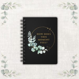 Puede incluir: Cuaderno negro con espiral con el texto "HOW DOES THIS MOMENT FEEL?" en dorado. La portada presenta un círculo dorado y un diseño de eucalipto estilo acuarela en tonos verdes y azules.