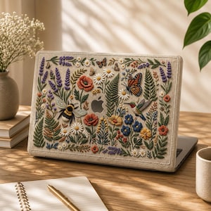 MacBook case with floral embroidery effect design featuring wildflowers, lavender, poppies, ferns, daisies, monarch butterfly, hummingbird, and bumblebee on warm linen-texture background. Shown on sunlit wooden desk with open notebook, gold pen, ceramic mug, and white wildflower stems. Soft natural light creates warm airy atmosphere. Beautiful botanical gift for cottagecore lovers, botanical art enthusiasts, and women who love nature-inspired workspace aesthetics.