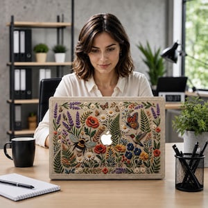 Woman working on MacBook with floral embroidery effect case featuring wildflowers, lavender, poppies, monarch butterfly, hummingbird, and bumblebee on linen-texture background. Shown in modern office workspace with industrial shelving, plants, black coffee mug, and notebook. Botanical cottagecore design brings nature-inspired aesthetic to professional work environment. Perfect botanical MacBook case for women who want to express their style at work.