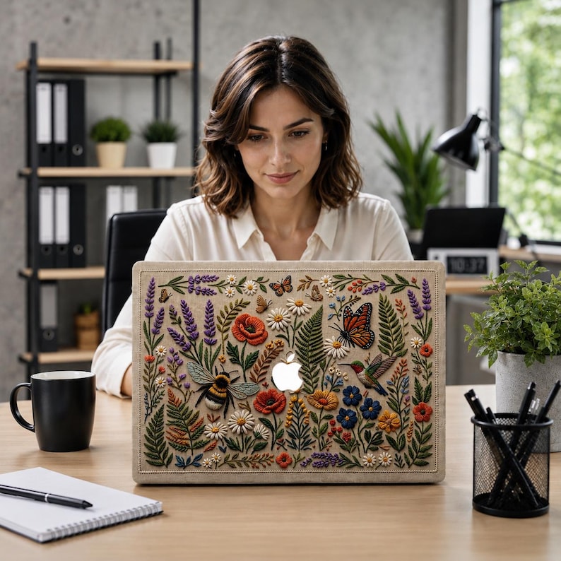 Woman working on MacBook with floral embroidery effect case featuring wildflowers, lavender, poppies, monarch butterfly, hummingbird, and bumblebee on linen-texture background. Shown in modern office workspace with industrial shelving, plants, black coffee mug, and notebook. Botanical cottagecore design brings nature-inspired aesthetic to professional work environment. Perfect botanical MacBook case for women who want to express their style at work.