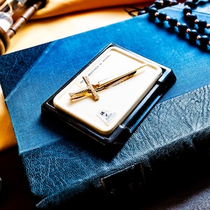 May include: A gold-toned sword-shaped tie bar resting on a rectangular white display card with the text MOTHER OF PEARL. The tie bar features a detailed hilt and a pointed blade, presented in a black plastic case set against a blue textured book.