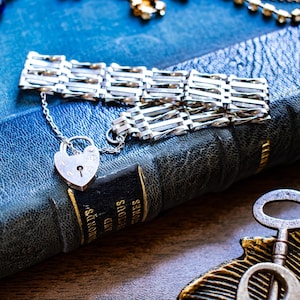 Könnte beinhalten: Ein silbernes Armband mit einer Reihe rechteckiger, durchbrochener Glieder, die über eine feine Kette mit einem herzförmigen Vorhängeschloss-Anhänger verbunden sind. Das Armband liegt auf dem Einband eines dunkelblauen, strukturierten Buches.