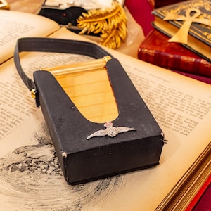 May include: A rectangular black case with a gold-toned metal center panel and a black strap handle. A silver-colored metal emblem featuring a pair of wings and a crown is attached to the front of the black casing, which rests on an open book.