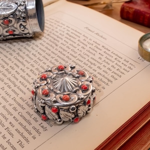 May include: A round silver box with a lid featuring a central sunburst pattern and small red coral inlays, surrounded by embossed leaf motifs. A matching silver cylindrical container with similar red coral and leaf details sits nearby.