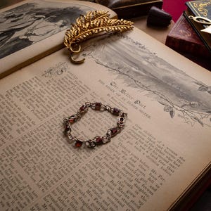 May include: A delicate silver-toned bracelet featuring small, dark red circular stones, resting on the open pages of an antique book. A gold-toned metal feather brooch lies nearby, alongside a small book with an ankh symbol on the cover.