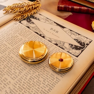 May include: Two circular gold-toned metal objects with smooth, reflective surfaces rest on an open antique book. One object features a small, embedded red gemstone at its center. The book page displays the text BAB by Alice E. Waddington.