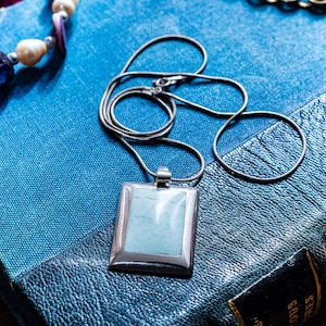 May include: A silver-toned metal necklace featuring a rectangular pendant with a smooth, light blue stone inlay. The pendant hangs from a thin, flexible snake chain, creating a clean and polished look with a simple, geometric design.