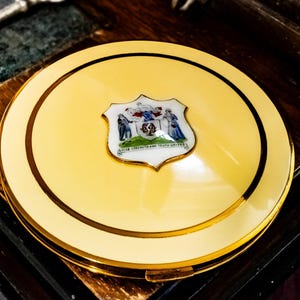 May include: A round, pale yellow compact mirror with a gold-toned metal rim and a central white shield-shaped emblem. The emblem features a detailed crest with two figures and the text "STRENGTH AND TRUTH UNITED" printed in black lettering.