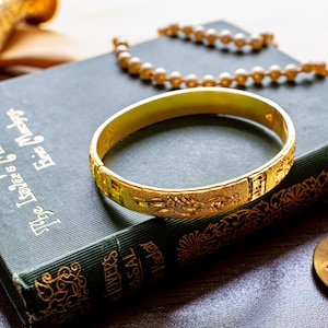 May include: A gold-toned metal bangle bracelet with an intricate textured pattern resting on a dark green hardcover book titled The Lover's Missal by Eric Mackay. The bracelet features a polished finish and a hinged clasp mechanism for closure.