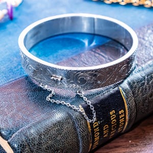 May include: A silver-toned metal bangle bracelet featuring an intricate engraved scrollwork pattern across its outer surface. The bracelet includes a hinged opening mechanism secured by a small metal safety chain attached to the sides of the band.