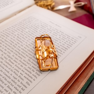 May include: A gold-toned metal ring featuring a rectangular frame design with multiple arched bands connected to a flat base. The ring is resting on the open pages of a book with printed text, showcasing a polished finish and a structured geometric shape.