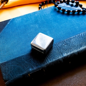 May include: A rectangular blue book with a textured cover sits beneath a small, polished silver-toned square box. Nearby are a wooden hourglass, a string of dark matte beads, a brass magnifying glass, and a small rectangular metal object.
