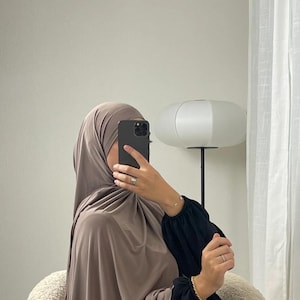 May include: A taupe-coloured hijab and matching long-sleeved garment featuring black sleeves with gathered cuffs. The fabric appears smooth and lightweight, draped over the head and shoulders, creating a layered look with contrasting black accents.