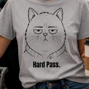 May include: A light gray t-shirt with a black graphic of a grumpy cat face and the words "Hard Pass." The person is wearing sunglasses and holding a coffee cup. The background shows a grocery store.