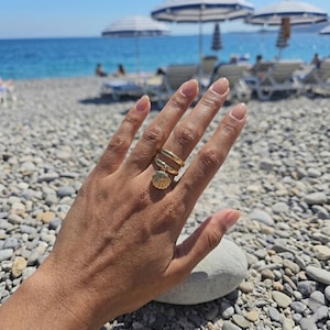 Puede incluir: Un anillo dorado con un dije de concha. El anillo tiene un diseño en espiral con un pequeño y detallado colgante de concha. La mano está enfocada, con una playa y el mar de fondo.