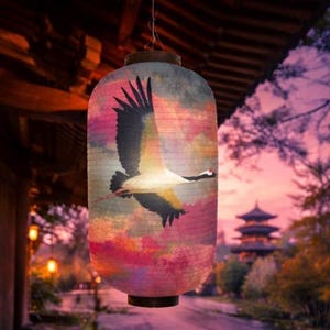 Puede incluir: Una linterna de papel con un diseño de grulla. La linterna presenta una colorida ilustración de una grulla en vuelo sobre un fondo de nubes rosas, moradas y azules. La linterna está colgada al aire libre, con una pagoda visible al fondo.